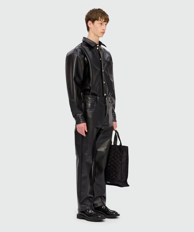 Male model wearing black long-sleeved shirt with structured faux leather pocket and matching pants, holding a tote bag.