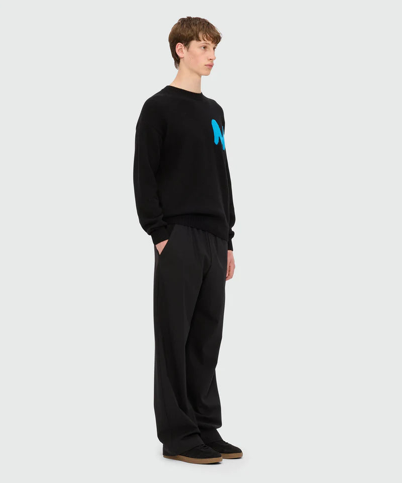 Black cashmere blend crew-neck sweater with TheMwave patch, model wearing size M/48, paired with loose black trousers.