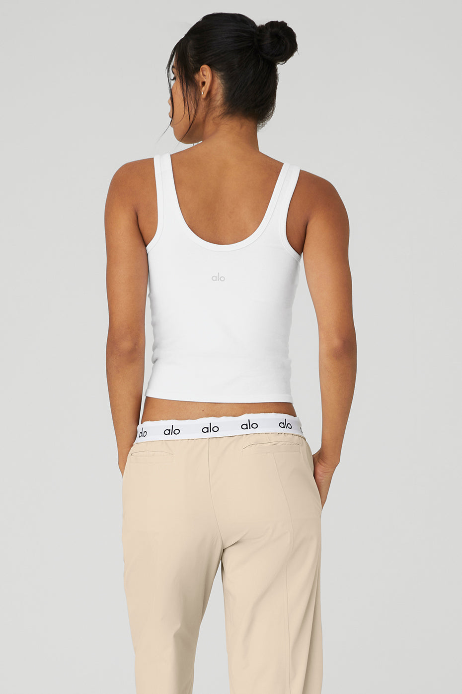 Back view of a woman wearing a white Seamless Chosen Tank with a deep scoop neckline and beige pants.