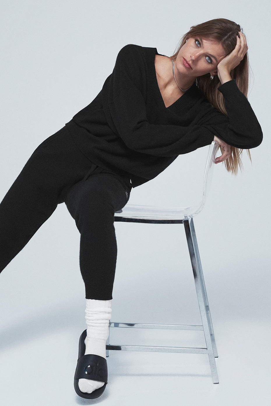 Model wearing Cashmere Jet Set V-Neck Pullover with dropped shoulders and V-neck sitting on a chair in black outfit.