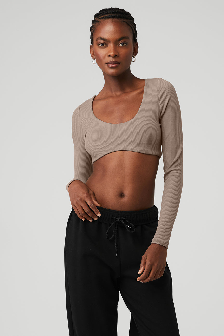 Model wearing a ribbed defined long sleeve bra in beige, showcasing a scoop neck and cropped design with flared pants.