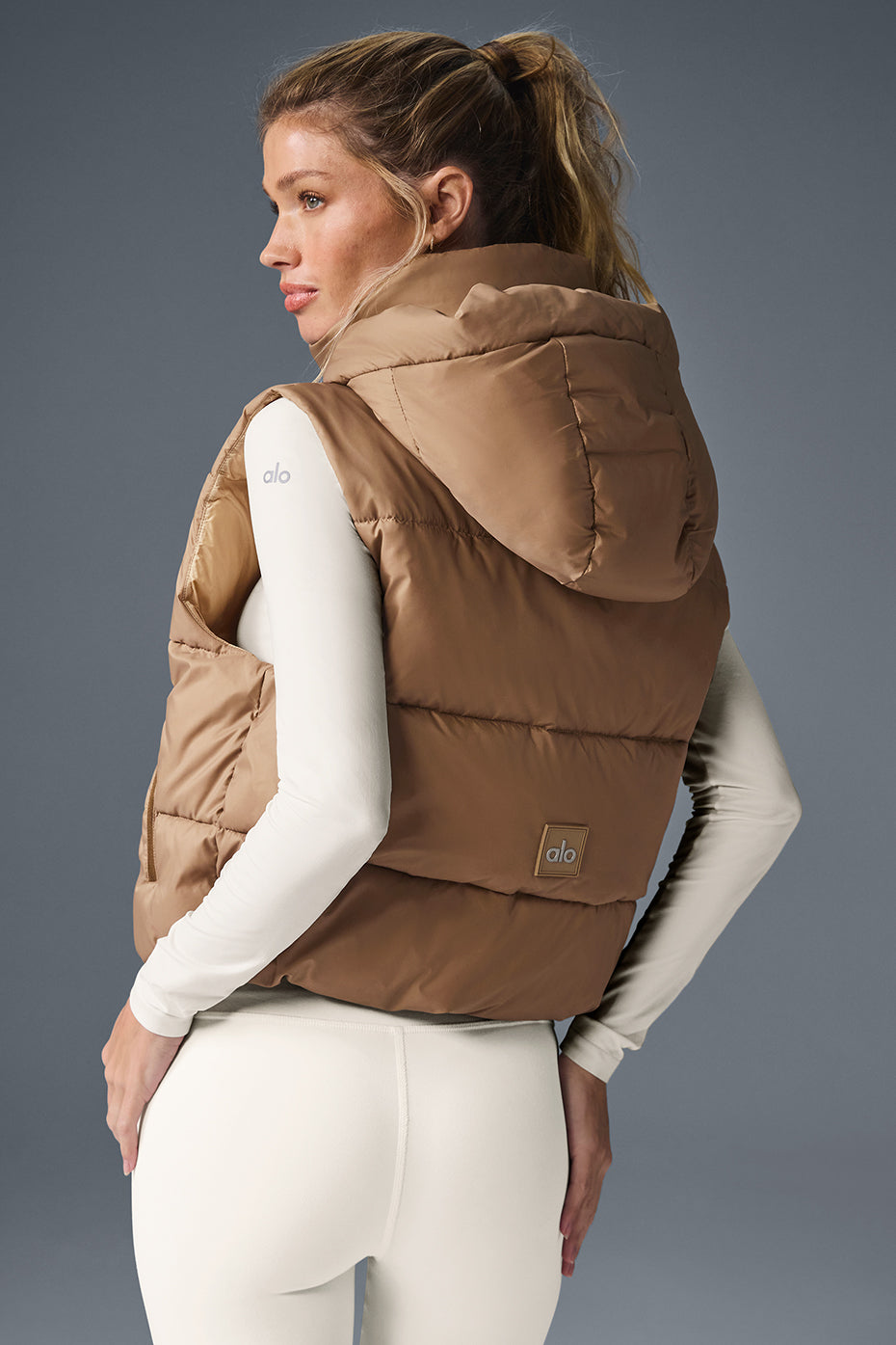Back view of a woman wearing the Gold Rush Puffer Vest in a cozy-luxe design with a hood and cinched waist.
