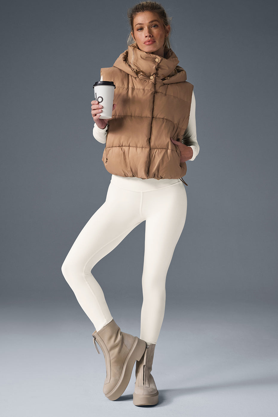 Model wearing Gold Rush Puffer Vest in beige with hood, paired with white leggings and holding a coffee cup.
