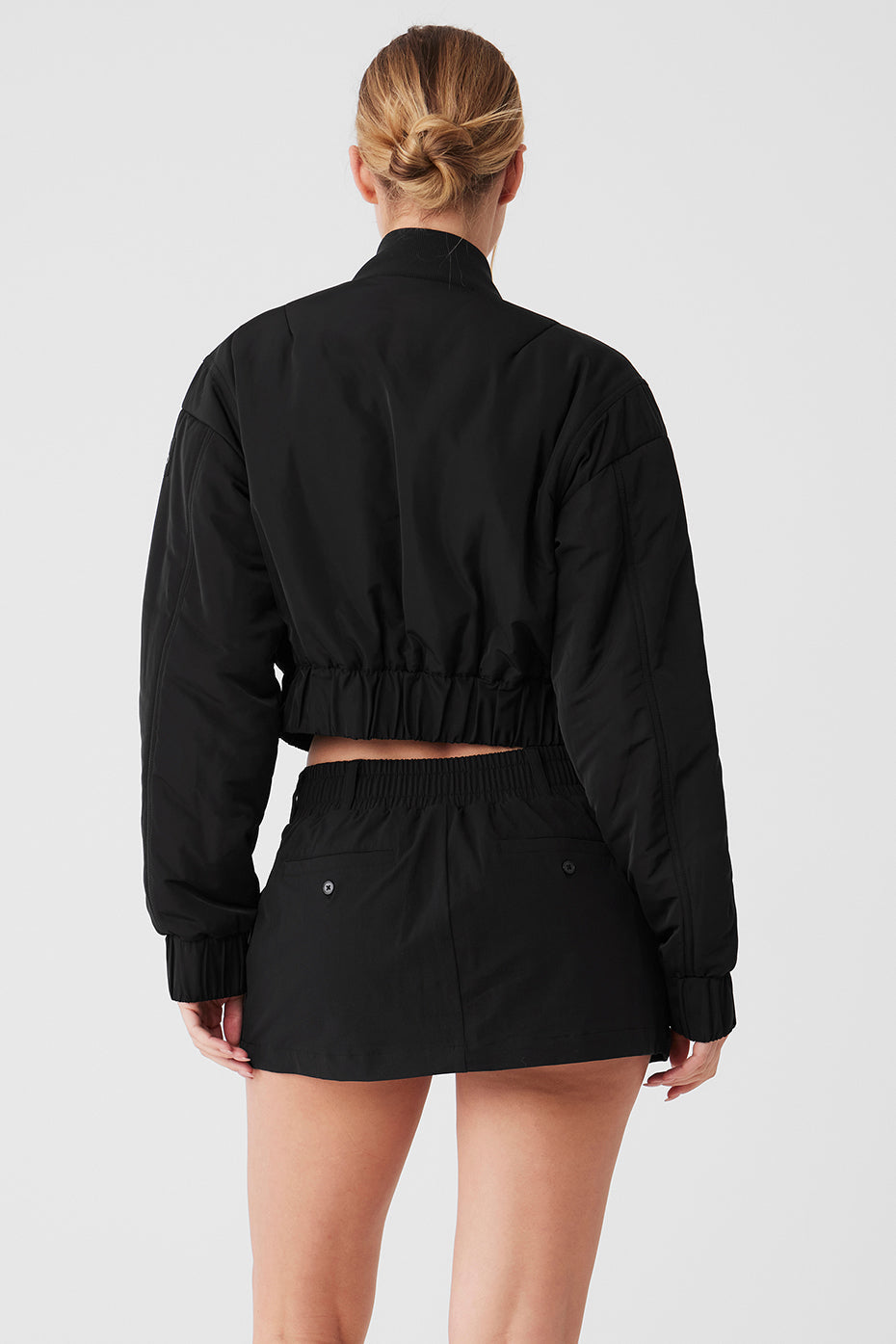 Back view of a woman wearing a cropped black bomber jacket with a ribbed collar and waistband.