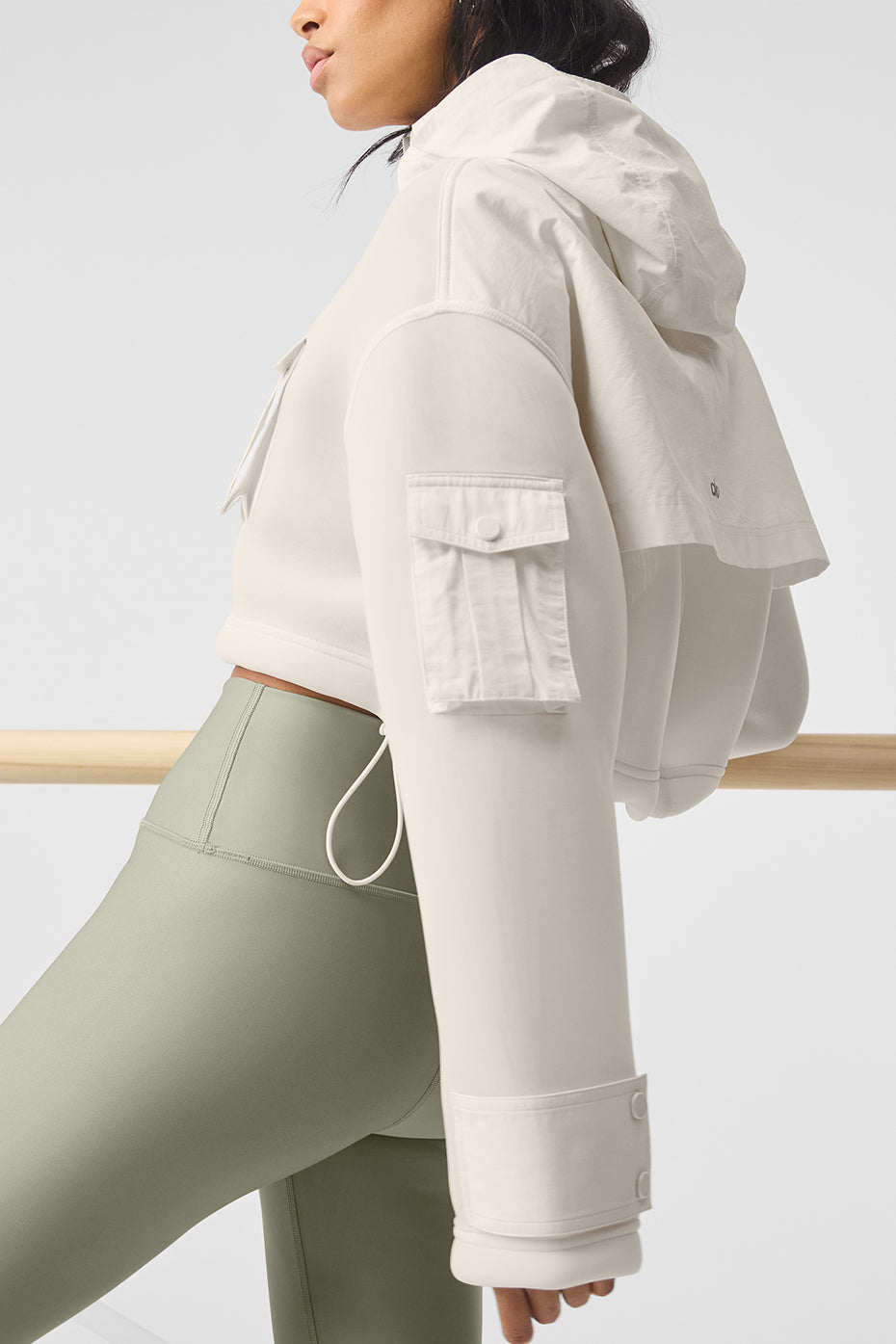 Stylish woman wearing a cropped Engage cargo parka jacket with hood and snap pockets, paired with green leggings.