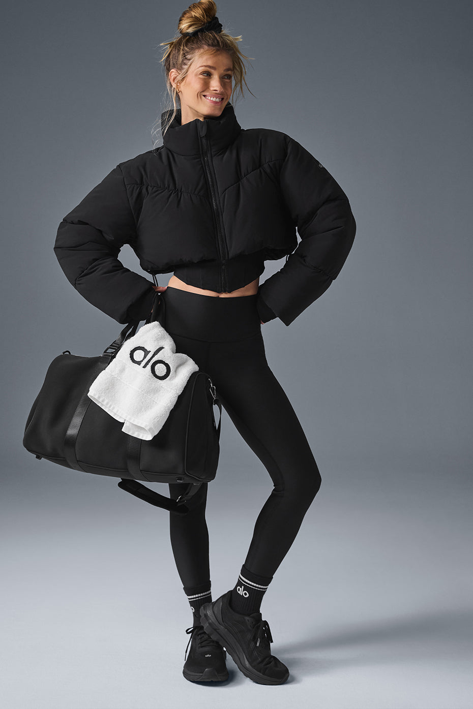 Model wearing Main Act Corset Puffer jacket with cropped silhouette and black leggings, holding a gym bag.