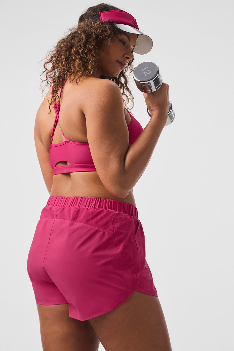 Woman in pink Playmaker Dolphin Shorts holds a dumbbell, showcasing athletic style and comfort during a workout.