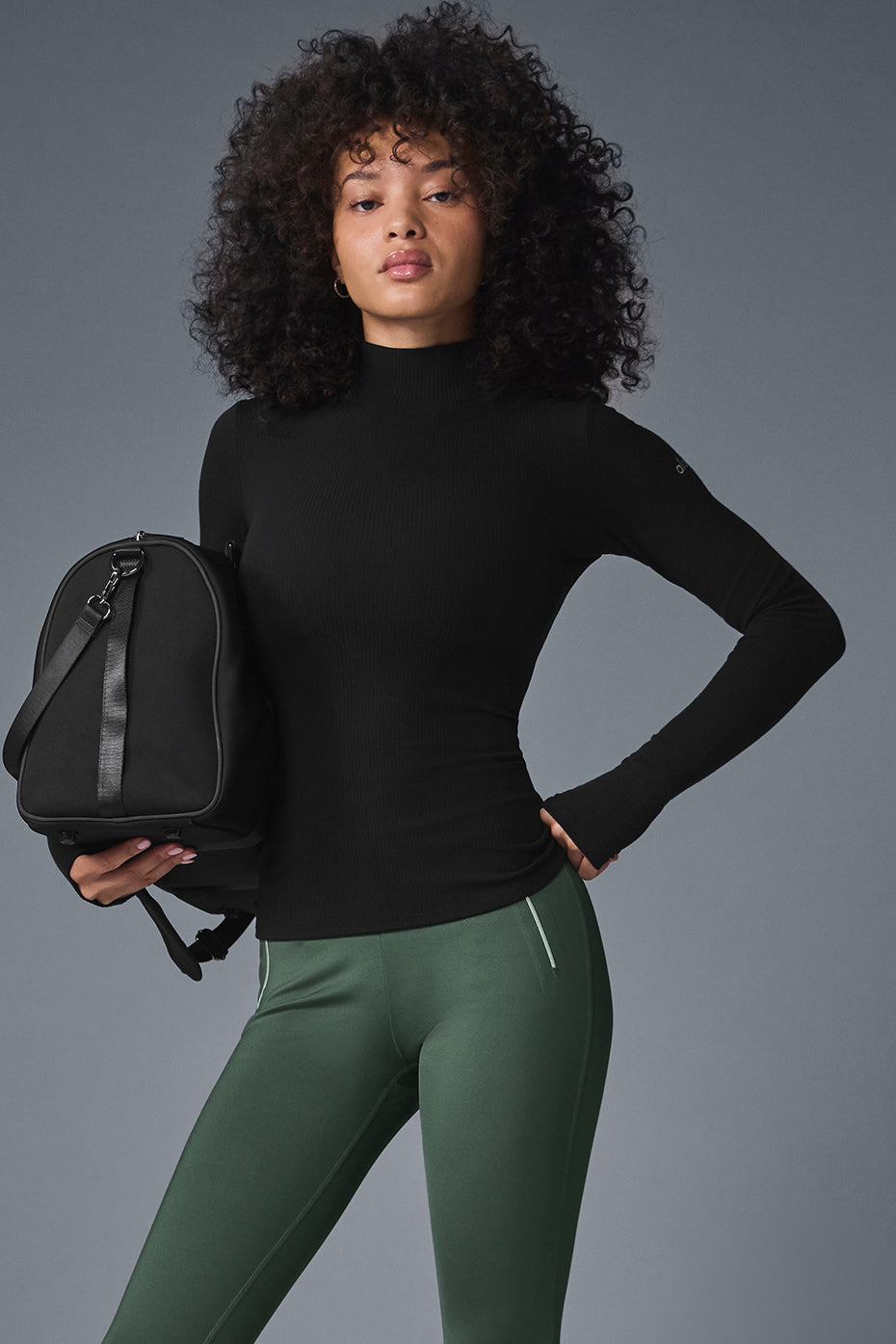 Model wearing Wellness Rib Mock Neck Long Sleeve top paired with green leggings and a black bag, showcasing a sleek and comfy look.