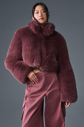 Opulent faux fur cropped jacket in rich burgundy color with stand collar and boxy shape, styled with wide-leg pants.
