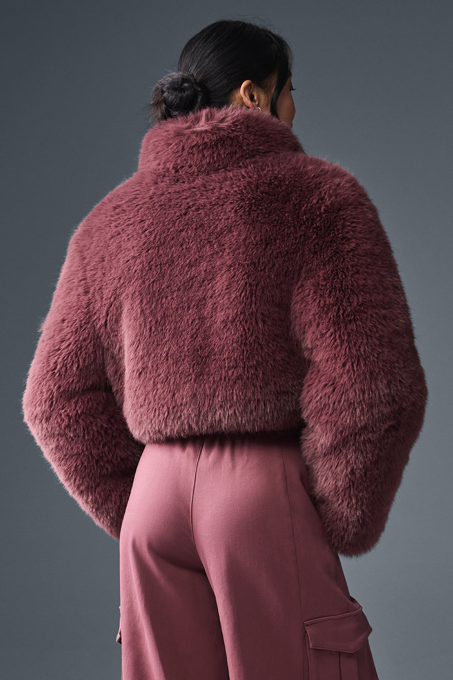 Back view of a woman wearing an opulent faux fur cropped jacket in a soft mauve color, styled with matching trousers.