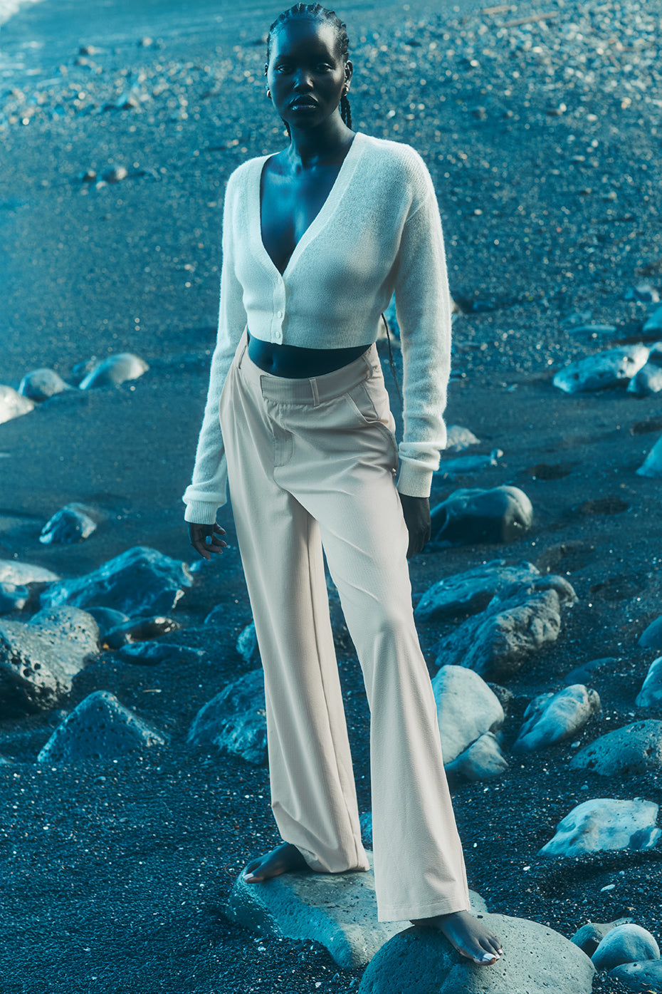 Model wearing High-Waist Dreamscape Trouser in lightweight fabric on rocky beach, showcasing stylish pleated design.