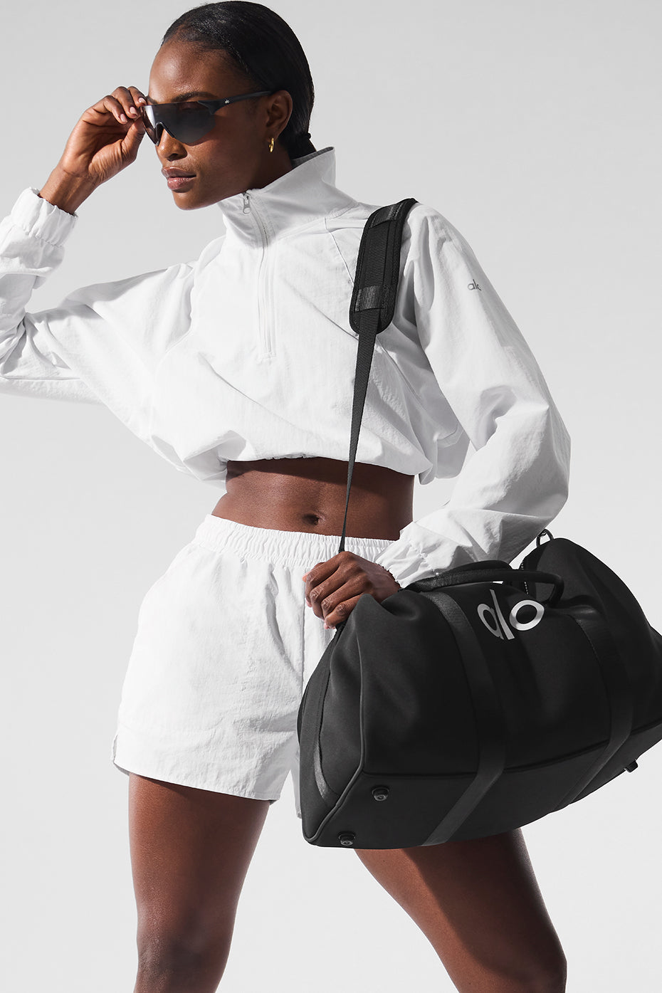 Model wearing a white cropped pullover and Alumni Shorts, carrying a black duffel bag for sports and outdoor activities.